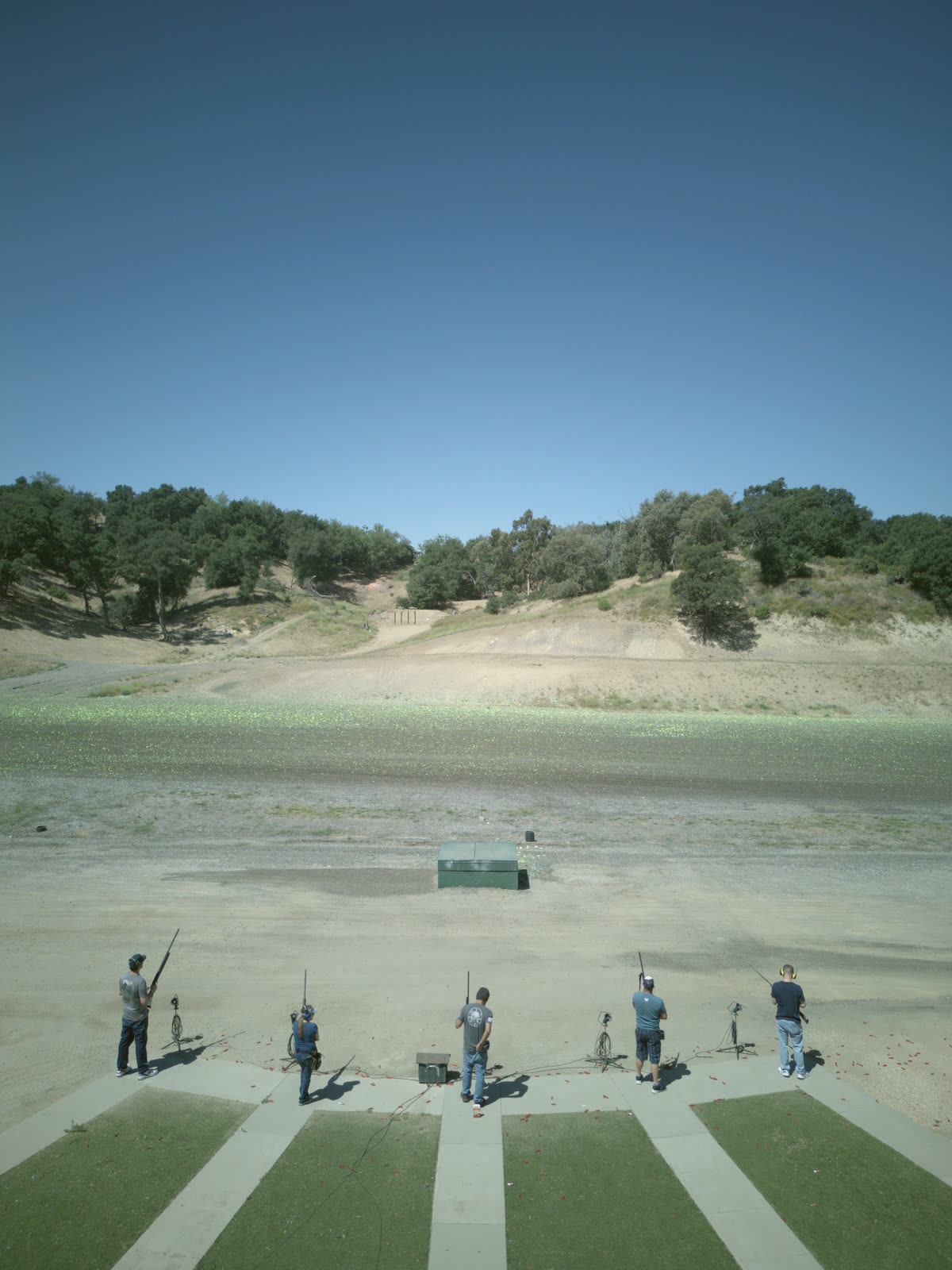 Five shooters on the pads of a trap field, the trap house ahead of them and the wooded hill beyond.